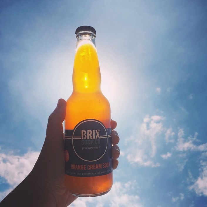 Hand holding Brix Orange Cream Soda with blue sky in the background